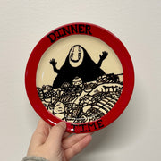 Round plate with a red rim featuring a black illustration of No Face and text, held against a white background.