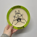Hand holding a ceramic plate with an illustration of Totoro and 'LUNCH TIME' text on a plain background