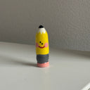 Small ceramic sculpture of a pencil with a cartoon smiley face on it.