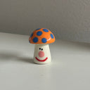 Tiny ceramic sculpture of an orange mushroom with blue dots and a cartoon smile on its stem.