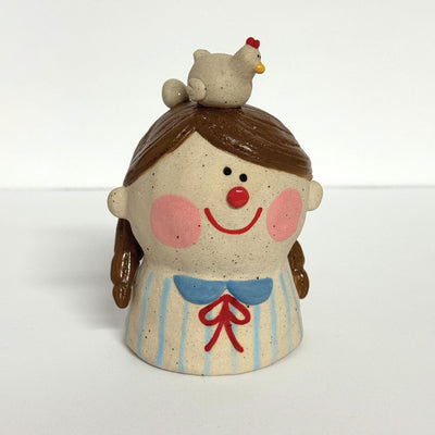 Ceramic sculpture of a girl's top half, without arms. She wears a striped shirt, smiles and has a chicken with an egg atop her head.