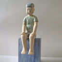 Carved wooden sculpture of a boy with a blank expression sitting on a platform with his hands on his knees and his legs dangling. He wears a t-shirt, shorts and a beanie. Colors are very muted cream, green and blue.