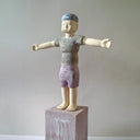 Carved wooden sculpture of a boy with a blank expression standing on a platform with both of his arms extended out as if stretching. He wears a t-shirt, shorts and a beanie. Colors are very muted cream, red, green and blue.