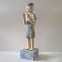Carved wooden sculpture of a boy with a blank expression standing and looking out at his hands, which he holds up to his face. He wears a t-shirt, shorts and a beanie. Colors are very mutes cream, blues and purple.