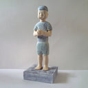 Carved wooden sculpture of a boy with a blank expression standing with his hands clasped together. He wears a t-shirt, shorts and a beanie. Colors are very mutes cream, blues and green.