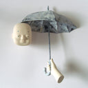 Carved wooden sculpture of a floating head. An open umbrella barely covers it, being held up by a floating hand. Colors are very muted cream and blue.