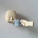 Carved wooden sculpture of a floating head, with a blue coffee cup being squished against its cheek by a floating hand. Colors are very muted cream and blue.