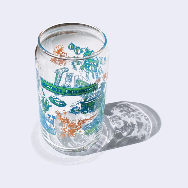 Glass cup with a flat base and slightly inward lip, it has printed design all over the cup. Various illustrations are within a color scheme of blue, green, orange. Illustrations include cherry blossoms, "Sawtelle Japantown" sign, a plant nursery and food.