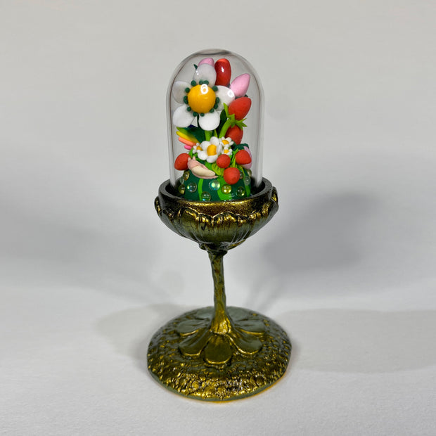 Small clay sculpture in a glass cloche atop a thin, elaborately decorated golden stand. Clay sculpture consists of several white flowers, greenery and many red and pink strawberries.