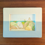 Watercolor illustration of a series of rocks, seen from underwater. 1 rock is shaped like a sleeping person's head. Around the scene are sea leaves. Piece if framed in half white, half blue frame.