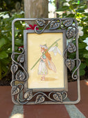 Illustration of a small flower fairy, standing in a side profile view and holding a large white flower on its stem. Piece is in an ornate metal leaf frame.