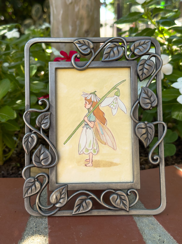 Illustration of a small flower fairy, standing in a side profile view and holding a large white flower on its stem. Piece is in an ornate metal leaf frame.