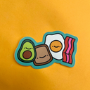 Die cut sticker of a group of brunch items in a cartoon art style: avocado, toast, egg and a slice of bacon.