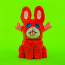 Vinyl figure of a red jackalope, sitting on the ground with hollowed blue eyes and pink small antlers.