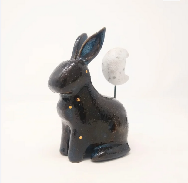 Ceramic sculpture of a black and blue bunny without any facial features and simplistic body features, sitting on its hind legs. Atop its back is a white speckled crescent moon.