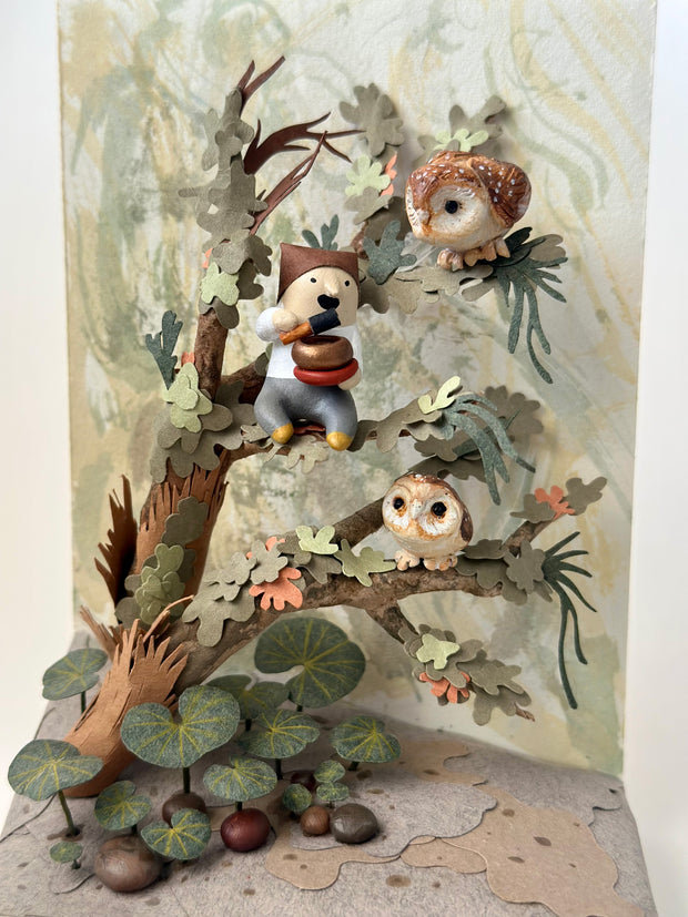 Mixed media sculpture of a person sitting on the branch of a tree, playing a sound bowl with small owls around them.