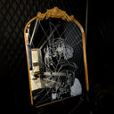 White engraved imagery on the surface of a mirror with gold gilding on the border. Imagery is of a woman looking over her shoulder with a shattered effect, splicing her face up.