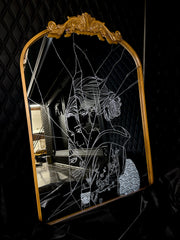 White engraved imagery on the surface of a mirror with gold gilding on the border. Imagery is of a woman looking over her shoulder with a shattered effect, splicing her face up.