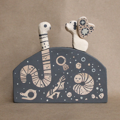 Wooden sculpture a mound with a worm and winged dog atop the ground, only partially painted with dark gray. Otherwise, natural wood color is exposed.  The mound is painted to show an x-ray of under ground life, such as worms, decomposing nature, rocks and a diamond ring. 