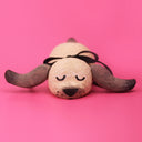 A handmade wooden dog sculpture with a natural light wood finish, dark brown painted ears and nose, and a black ribbon tail, lying on a solid pink background.