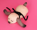 A handmade wooden dog sculpture with a natural light wood finish, dark brown painted ears and nose, and a black ribbon tail, lying on a solid pink background.