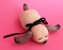 A handmade wooden dog sculpture with a natural light wood finish, dark brown painted ears and nose, and a black ribbon tail, lying on a solid pink background.