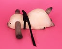 A handmade wooden dog sculpture with a natural light wood finish, dark brown painted ears and nose, and a black ribbon tail, lying on a solid pink background.