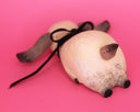 A handmade wooden dog sculpture with a natural light wood finish, dark brown painted ears and nose, and a black ribbon tail, lying on a solid pink background.