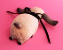 A handmade wooden dog sculpture with a natural light wood finish, dark brown painted ears and nose, and a black ribbon tail, lying on a solid pink background.