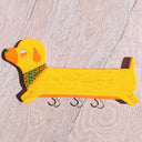 Die cut flat wooden sculpture of a yellow wiener dog, with a very long body and tiny legs. It has an orange snout, ear, and eyes and wears a floral green and orang bandana. It has hooks hanging from the bottom.