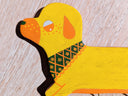 Die cut flat wooden sculpture of a yellow wiener dog, with a very long body and tiny legs. It has an orange snout, ear, and eyes and wears a floral green and orang bandana. It has hooks hanging from the bottom.