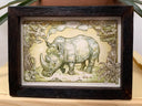A vintage-style illustration showing a rhinoceros at center, framed by ornate decorative foliage and scrollwork in muted beige, olive green, and brown tones on a light background.