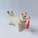 White ceramic sculpture of a dog like creature with a tassel hanging from its mouth and 2 tails. It has a necklace made of fangs as its collar.