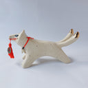 White ceramic sculpture of a dog like creature with a tassel hanging from its mouth and 2 tails. It has a necklace made of fangs as its collar.