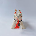 White ceramic sculpture of a dog like creature with a tassel hanging from its mouth and 2 tails. It has a necklace made of fangs as its collar.