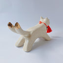 White ceramic sculpture of a dog like creature with a tassel hanging from its mouth and 2 tails. It has a necklace made of fangs as its collar.