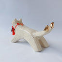 White ceramic sculpture of a dog like creature with a tassel hanging from its mouth and 2 tails. It has a necklace made of fangs as its collar.