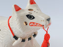 White ceramic sculpture of a dog like creature with a tassel hanging from its mouth and 2 tails. It has a necklace made of fangs as its collar.