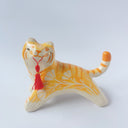 Glossy ceramic sculpture of an orange and white tiger with gold detailing, holding a red fabric tassel in its mouth.