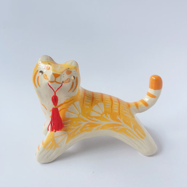 Glossy ceramic sculpture of an orange and white tiger with gold detailing, holding a red fabric tassel in its mouth.
