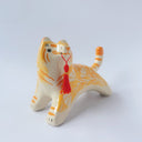 Glossy ceramic sculpture of an orange and white tiger with gold detailing, holding a red fabric tassel in its mouth.