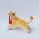 Glossy ceramic sculpture of an orange and white tiger with gold detailing, holding a red fabric tassel in its mouth.