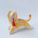 Glossy ceramic sculpture of an orange and white tiger with gold detailing, holding a red fabric tassel in its mouth.