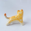Glossy ceramic sculpture of an orange and white tiger with gold detailing, holding a red fabric tassel in its mouth.