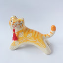 Glossy ceramic sculpture of an orange and white tiger with gold detailing, holding a red fabric tassel in its mouth.