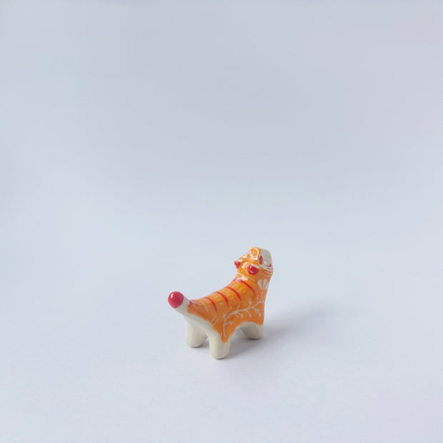 Very small ceramic sculpture of a tiger with a white underbelly and orange fur with red stripes, gold detailing and white carved poppy flowers. It is in a roaring stance.