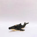 A small whale-shaped ceramic sculpture with a matte black finish and sgraffito beige details, including a beige underside and a gold circular accent on its side, set against a plain light gray background.