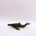 A small whale-shaped ceramic sculpture with a matte black finish and sgraffito beige details, including a beige underside and a gold circular accent on its side, set against a plain light gray background.