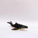 A small whale-shaped ceramic sculpture with a matte black finish and sgraffito beige details, including a beige underside and a gold circular accent on its side, set against a plain light gray background.