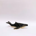 A small whale-shaped ceramic sculpture with a matte black finish and sgraffito beige details, including a beige underside and a gold circular accent on its side, set against a plain light gray background.